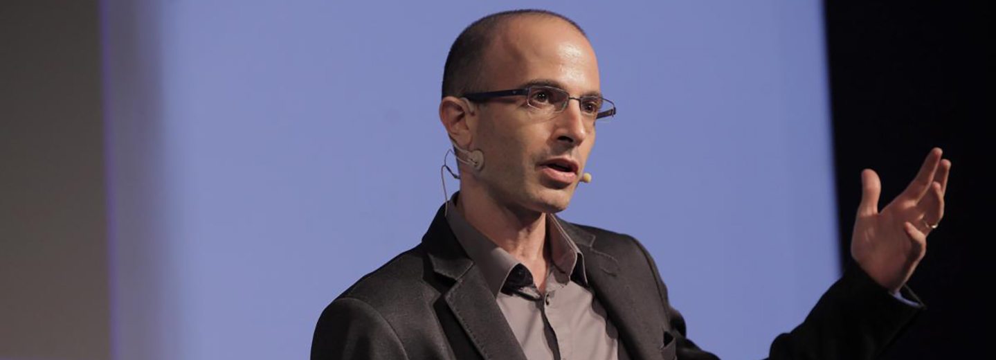 Yuval Noah Harari phát biểu trên sân khấu với micro cài tai, tay đưa lên minh hoạ khi trình bày